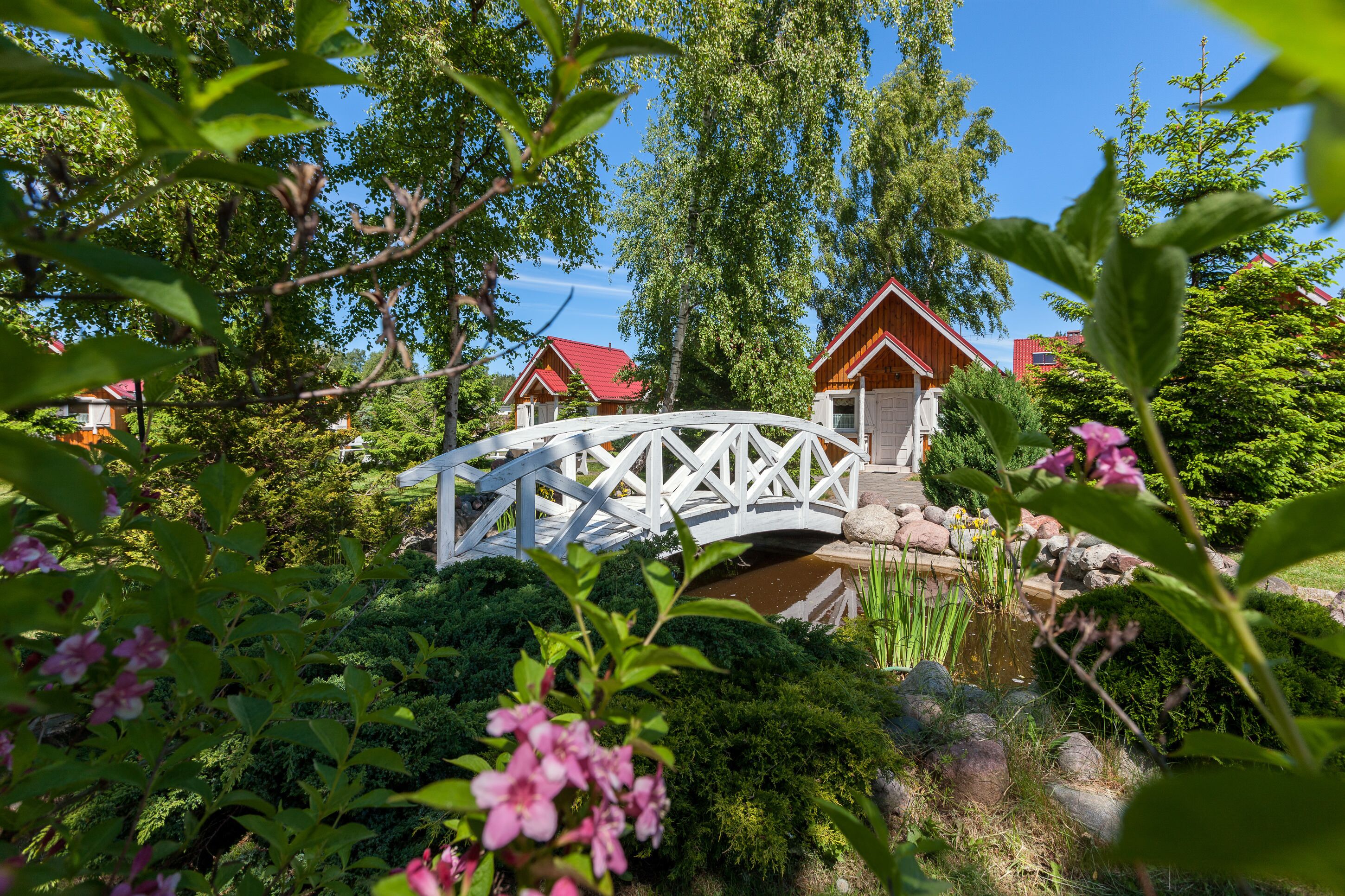 Eine weiße Brücke über einen Teich mit Holzhäusern im Hintergrund.