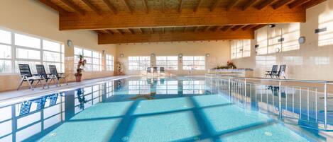 An indoor swimming pool with wooden beams, large windows, and sunlit areas.