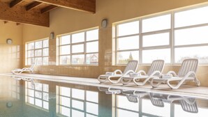 Piscina interior com paredes de vidro transparente, várias janelas e espreguiçadeiras à beira da piscina.