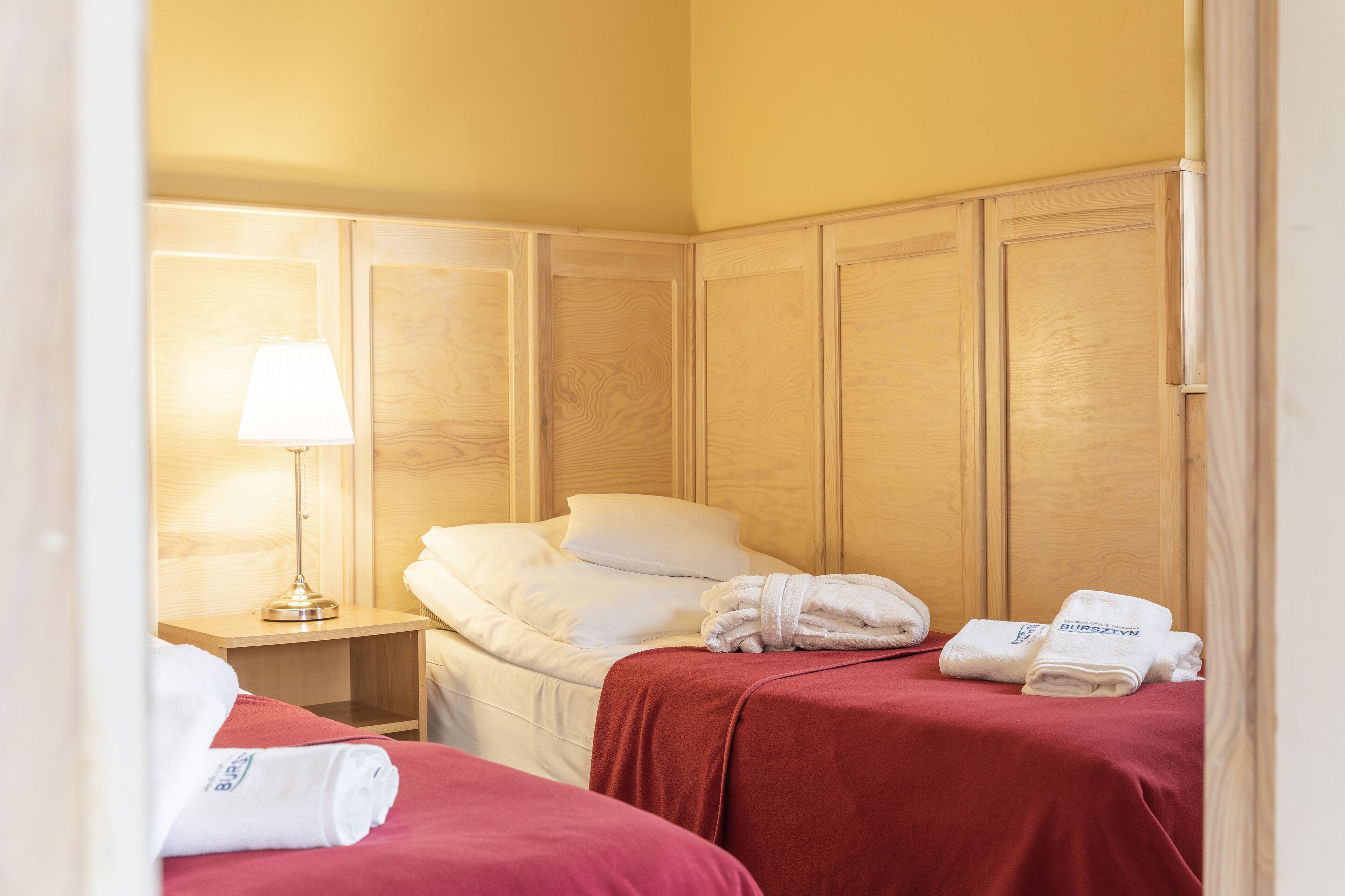 A hotel room with two beds, wooden paneling, a nightstand with a lamp, and towels on the beds.