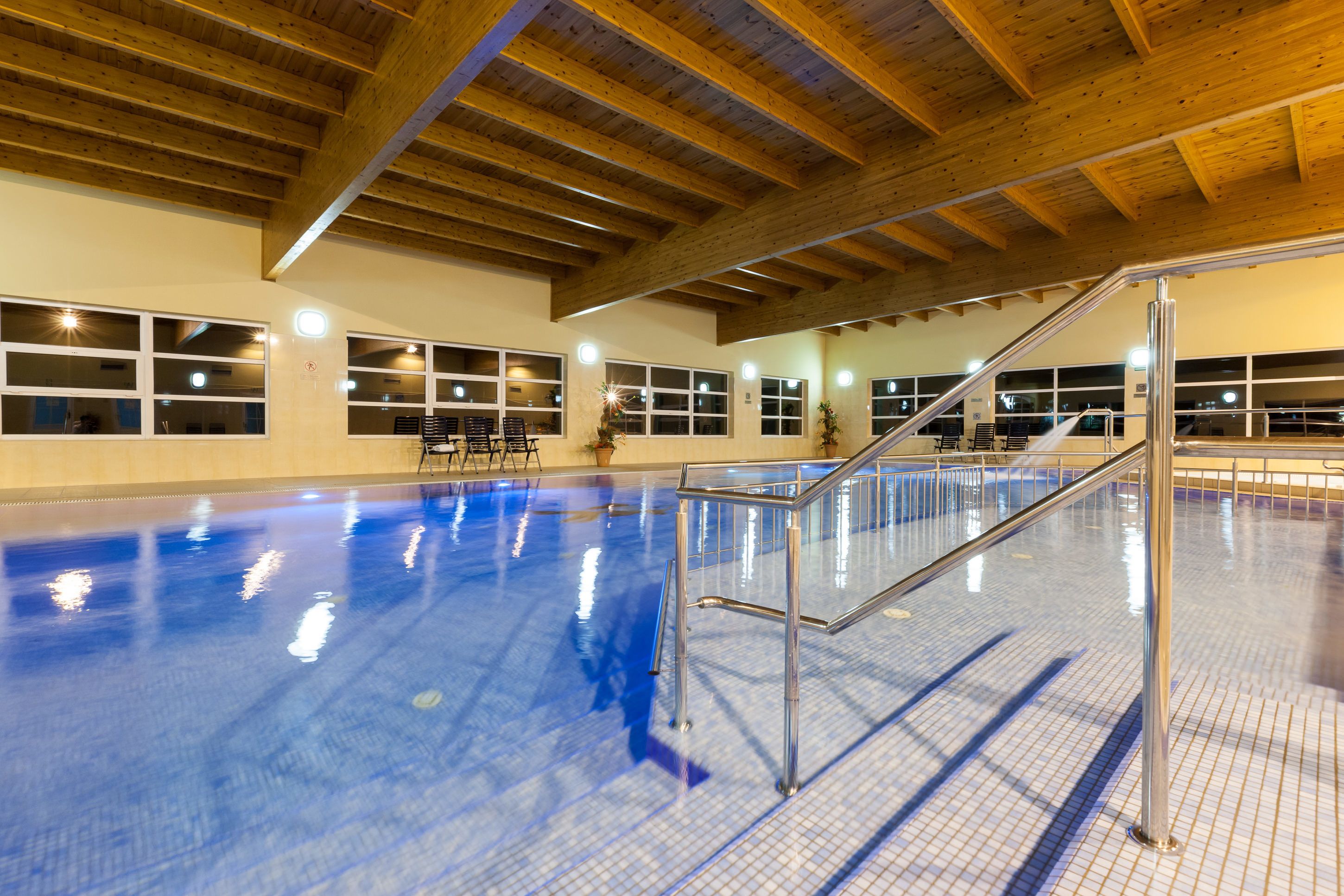An indoor swimming pool with a tiled floor, wooden ceiling beams, and a metal staircase leading to the upper level.