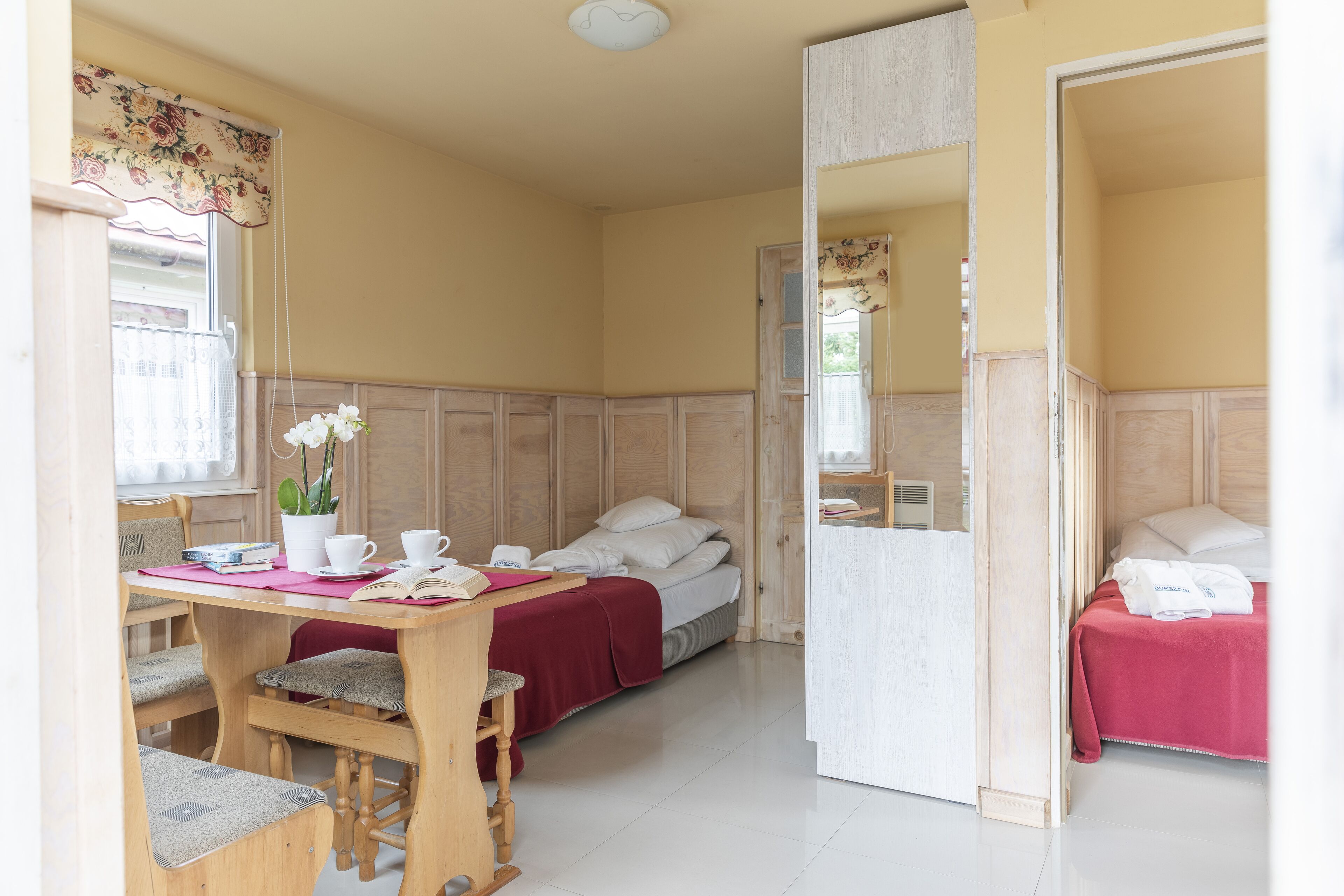 A room with a wooden desk, two beds, a mirror, and a window with curtains.