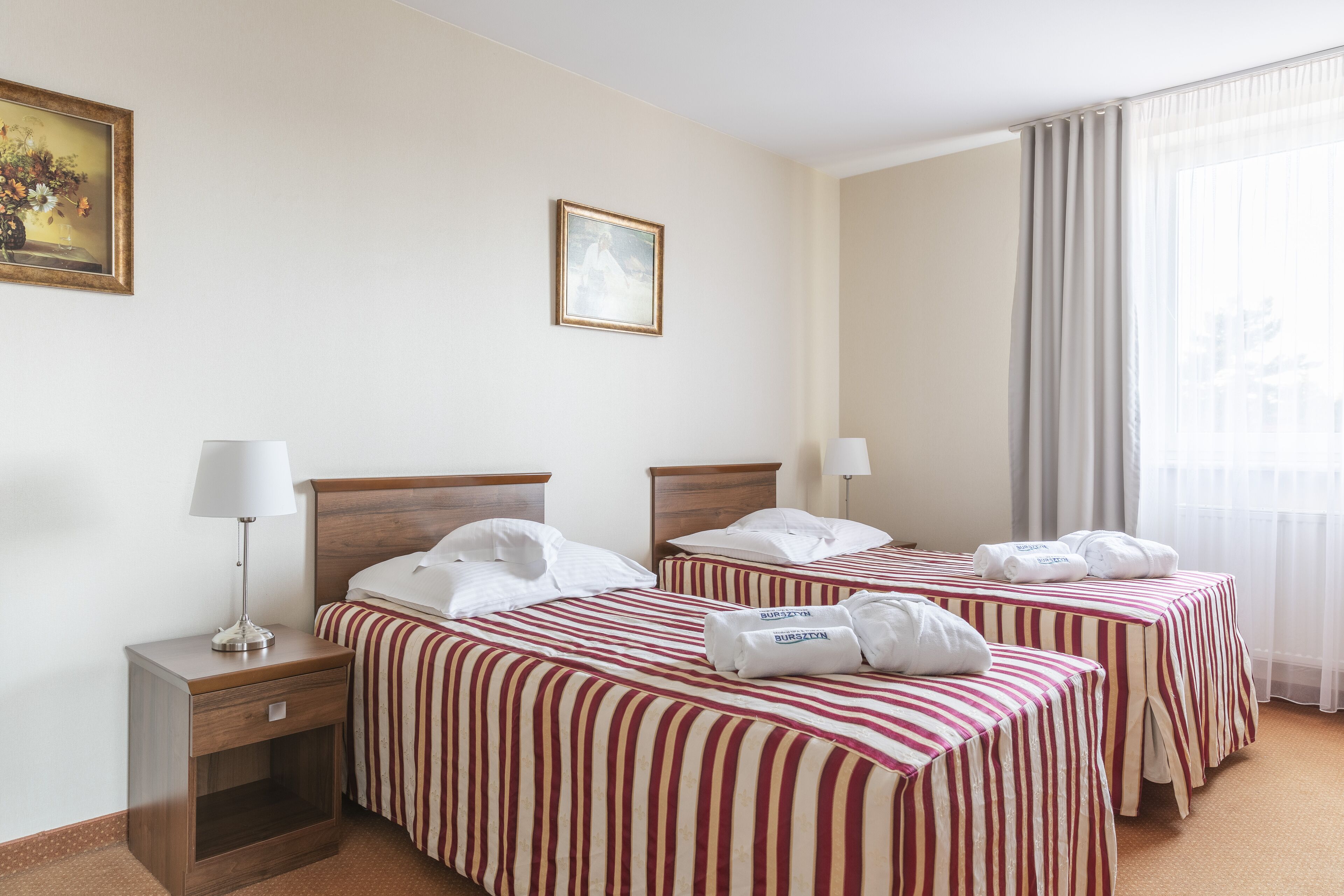 A hotel room with two beds, each with striped bedding and towels. There is a nightstand with a lamp, a framed picture on the wall, and a window with curtains.