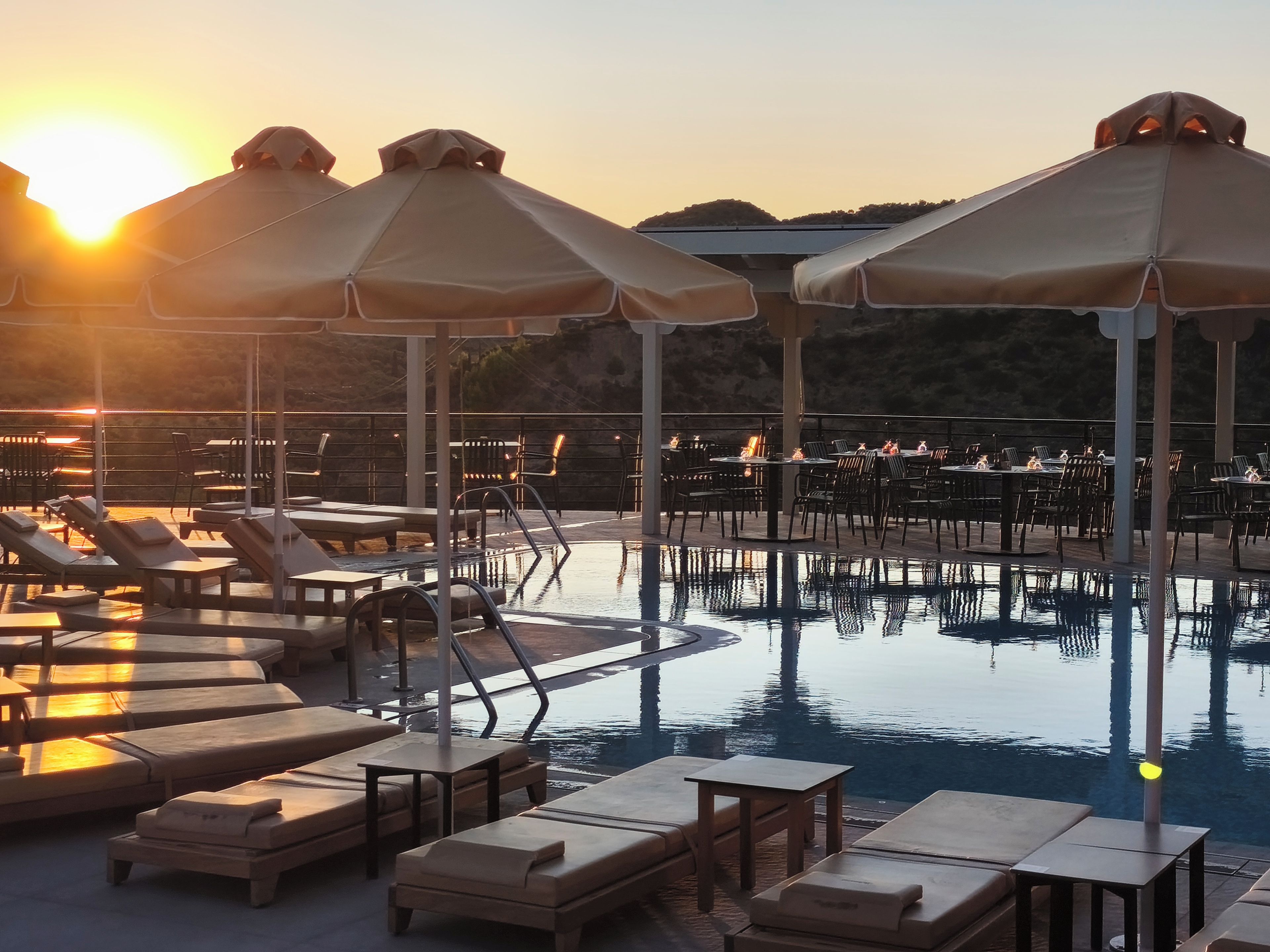Zona de piscina con tumbonas y sombrillas, rodeada de mesas y sillas, con vistas a un paisaje montañoso.