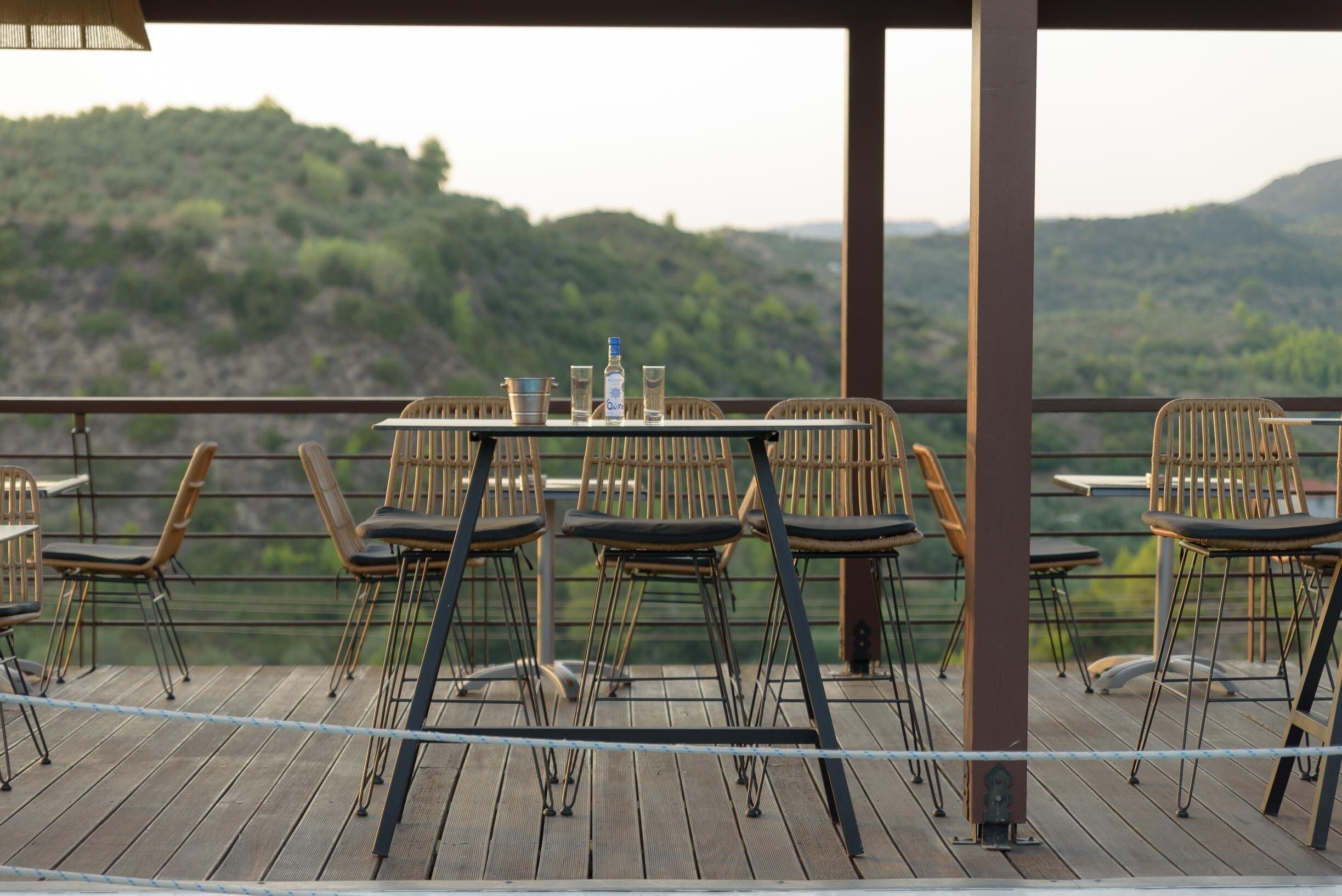 Una terraza de madera con asientos exteriores, una mesa con bebidas y una vista panorámica de las colinas.
