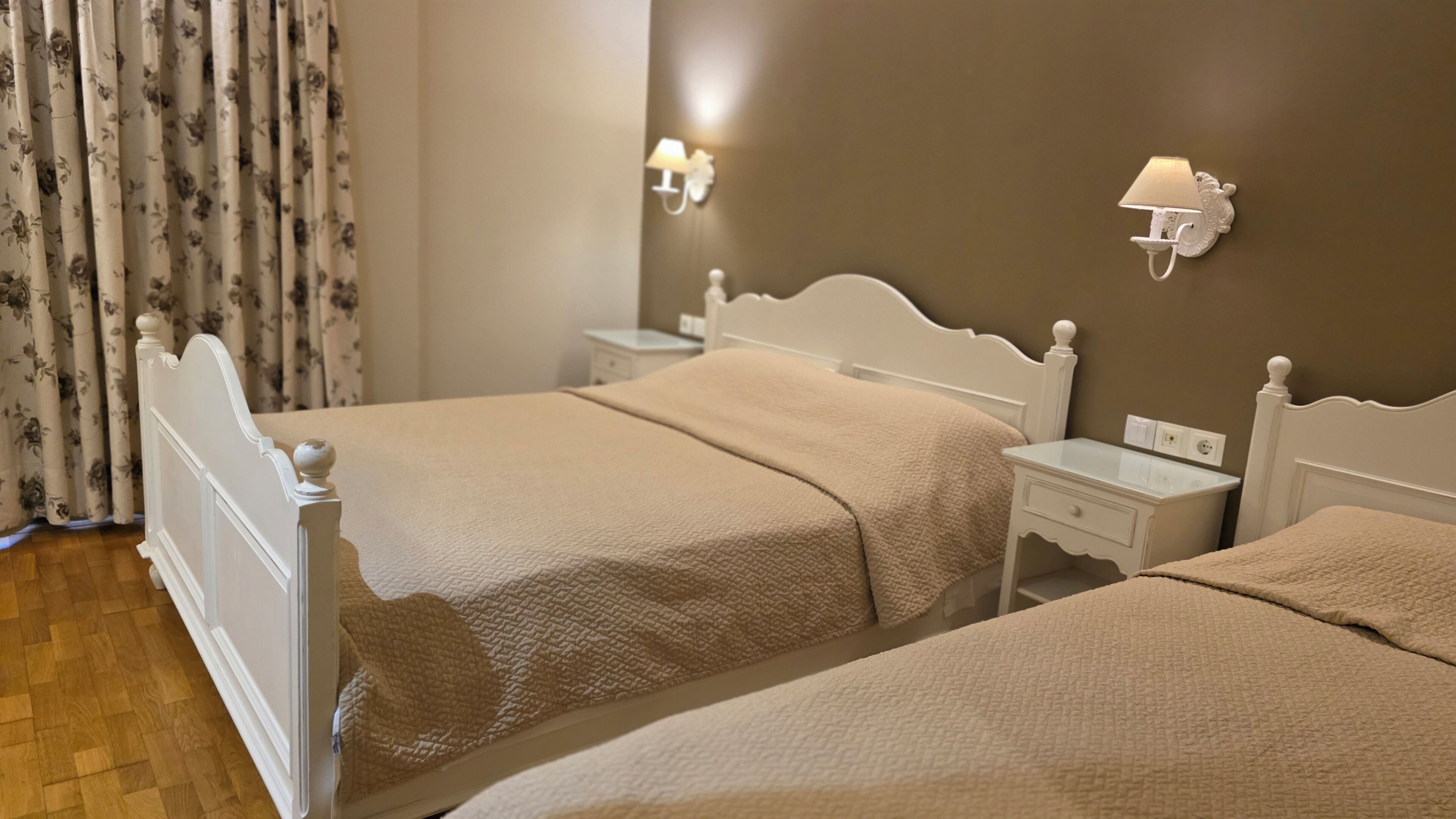 A hotel room with two beds, a nightstand, a lamp, and a curtain with a floral pattern.
