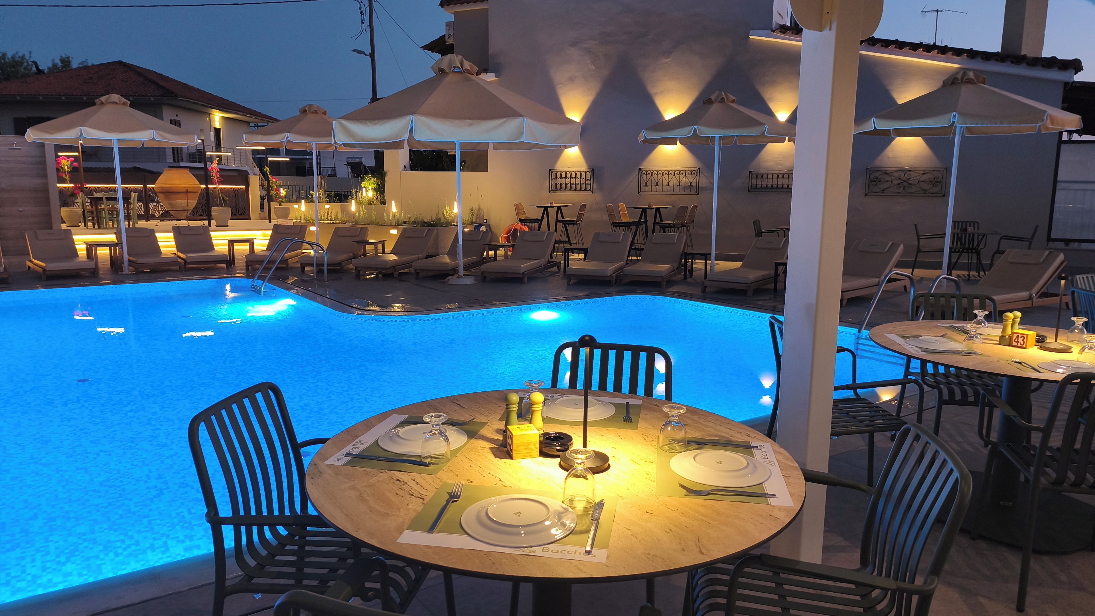 Un área de comedor junto a la piscina con mesas preparadas para dos personas, sillones de descanso y sombrillas.