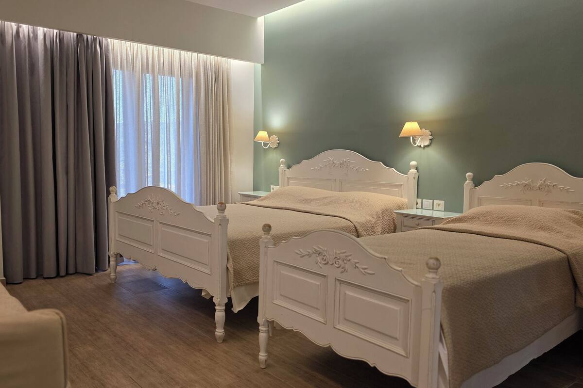 Un dormitorio con dos camas blancas, una ventana con cortinas, una mesita de noche con una lámpara y un radiador.