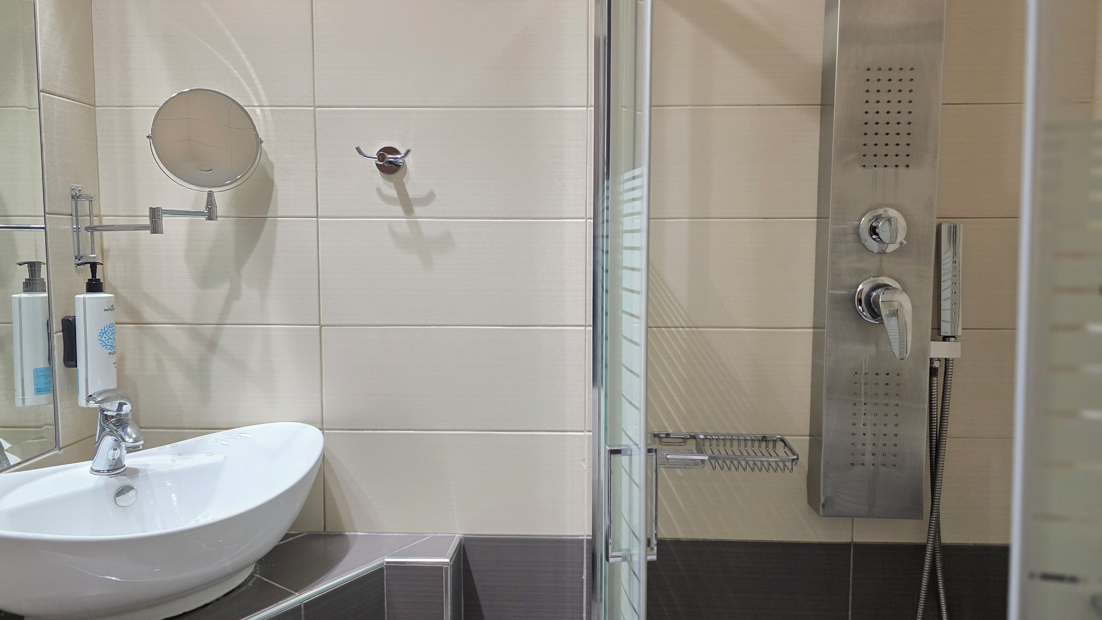 Un baño moderno con un lavamanos blanco, un espejo de mano, un cabezal de ducha de mano y una cabina de ducha con mampara de vidrio.