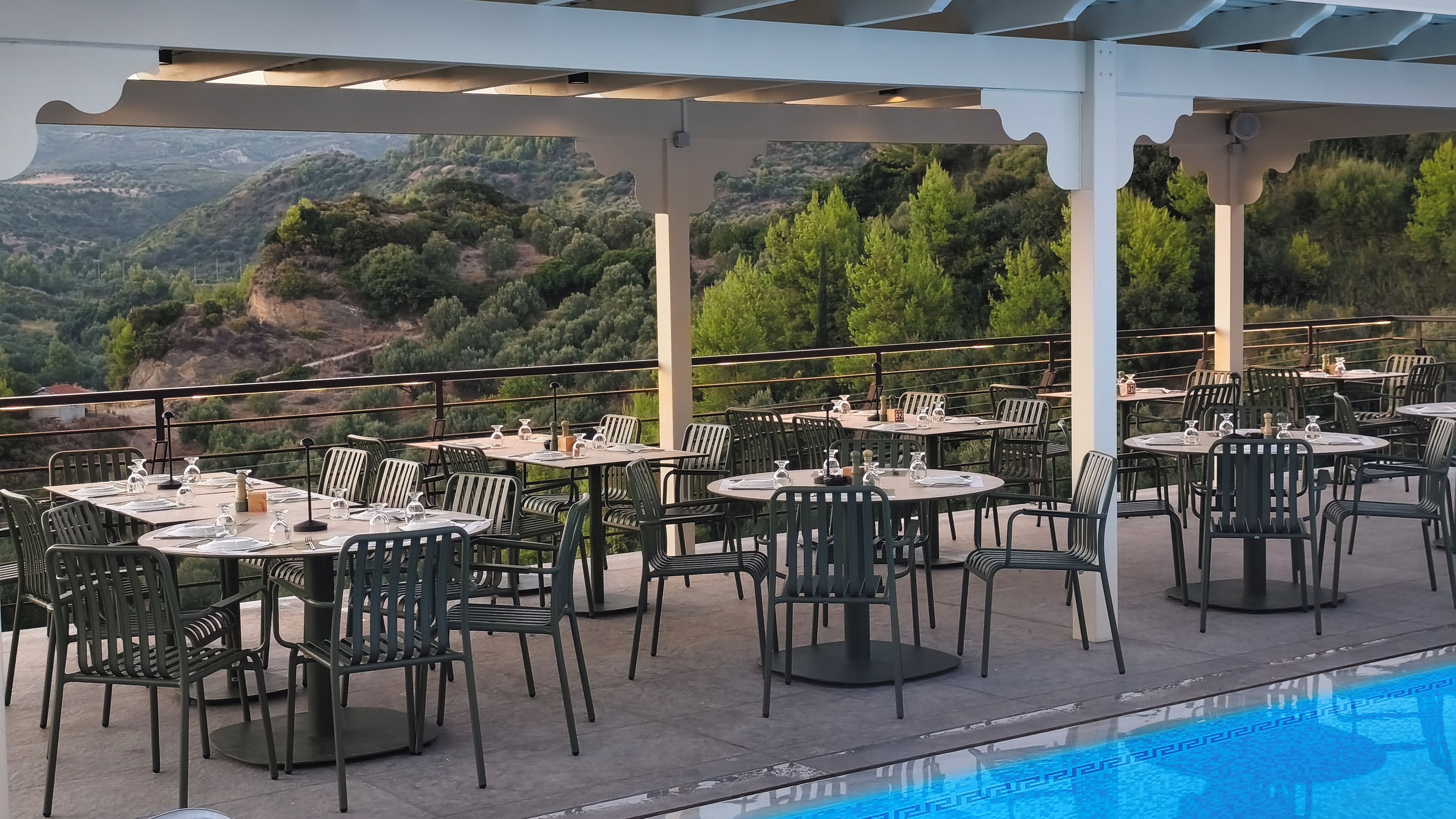 An outdoor dining area with tables and chairs, overlooking a pool and a scenic view of hills.