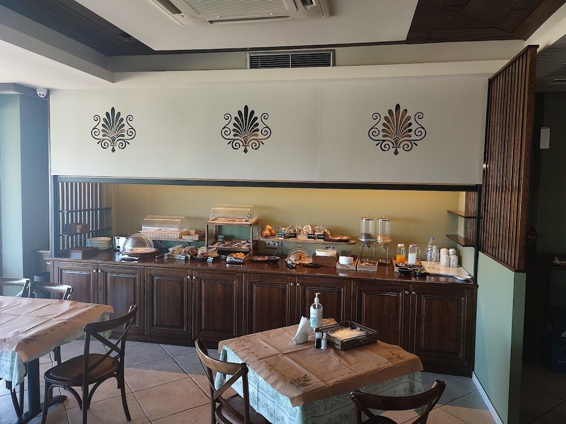 A breakfast bar with various food items and a coffee machine. There are wooden chairs and a table with a placemat.