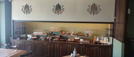 A breakfast bar with various food items and a coffee machine. There are wooden chairs and a table with a placemat.