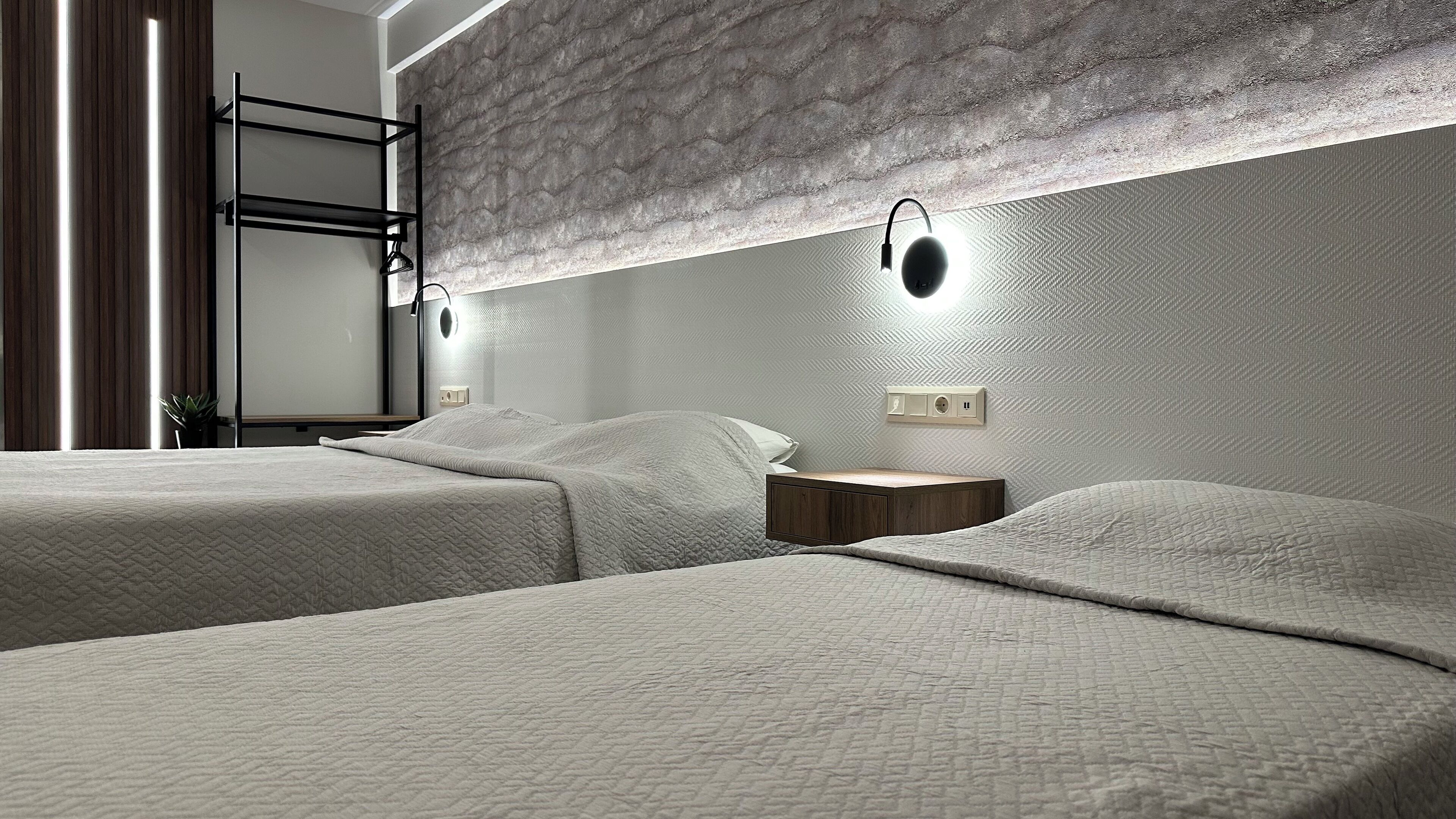 A modern hotel room with two beds, a wooden nightstand, a wall-mounted light, and a built-in shelf.