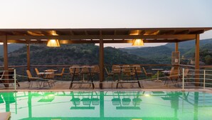 Zona de piscina exterior con sillas y mesas bajo una pérgola, rodeada por un paisaje montañoso.