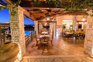 An outdoor dining area with stone walls and wooden beams, featuring tables and chairs arranged for guests. - Bacchus (Archaia Olympia)