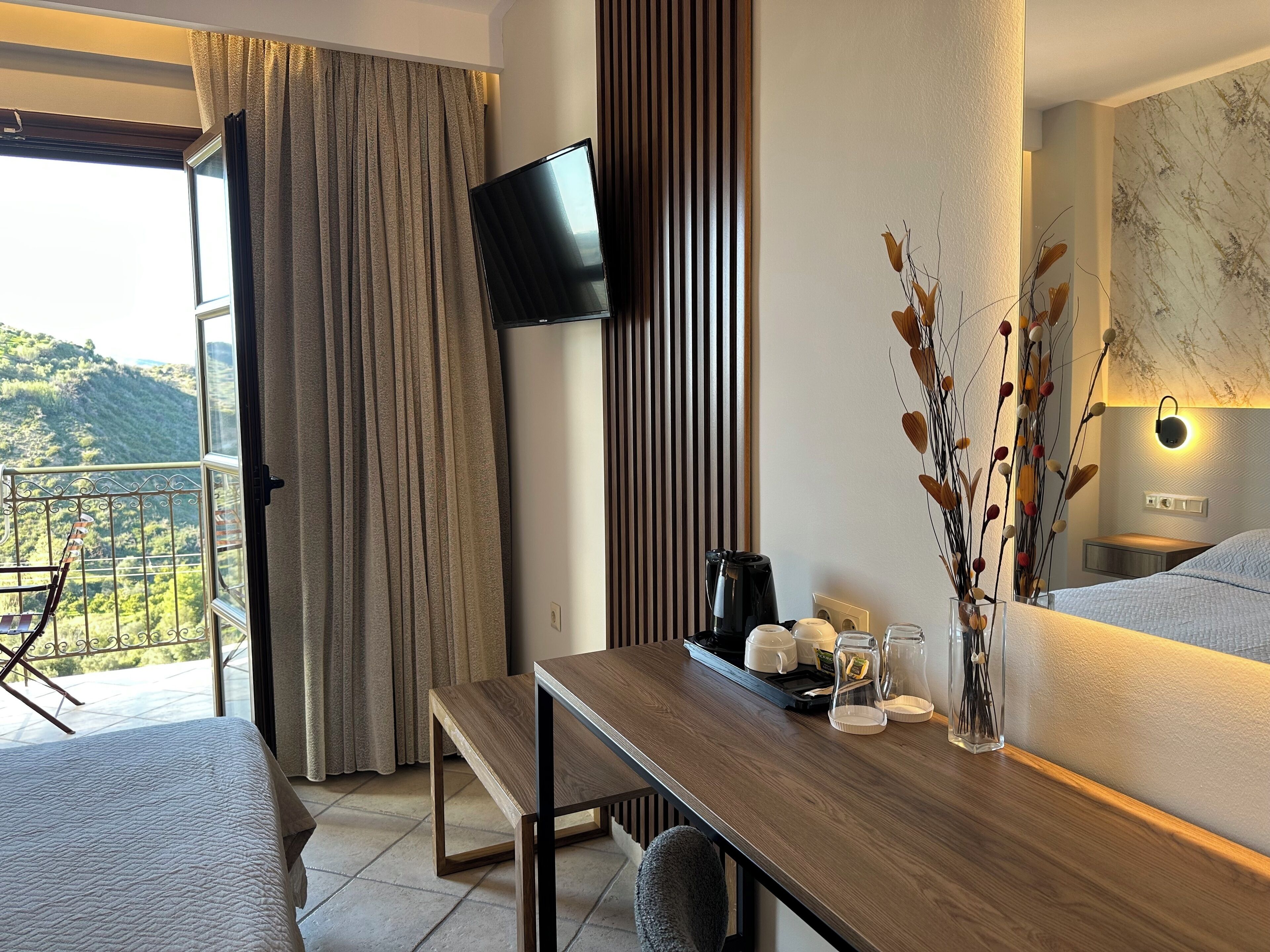 A hotel room with a wooden desk, a flat-screen TV mounted on the wall, a balcony with a view of the hills, and a bed with a white quilt.