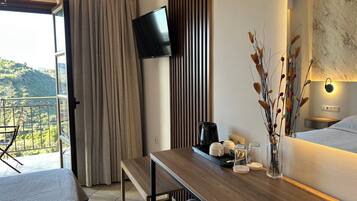 A hotel room with a wooden desk, a flat-screen TV mounted on the wall, a balcony with a view of the hills, and a bed with a white quilt.