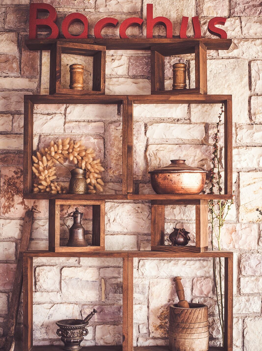Una estantería de madera rústica con diversos utensilios de cocina y objetos decorativos frente a un fondo de pared de piedra.