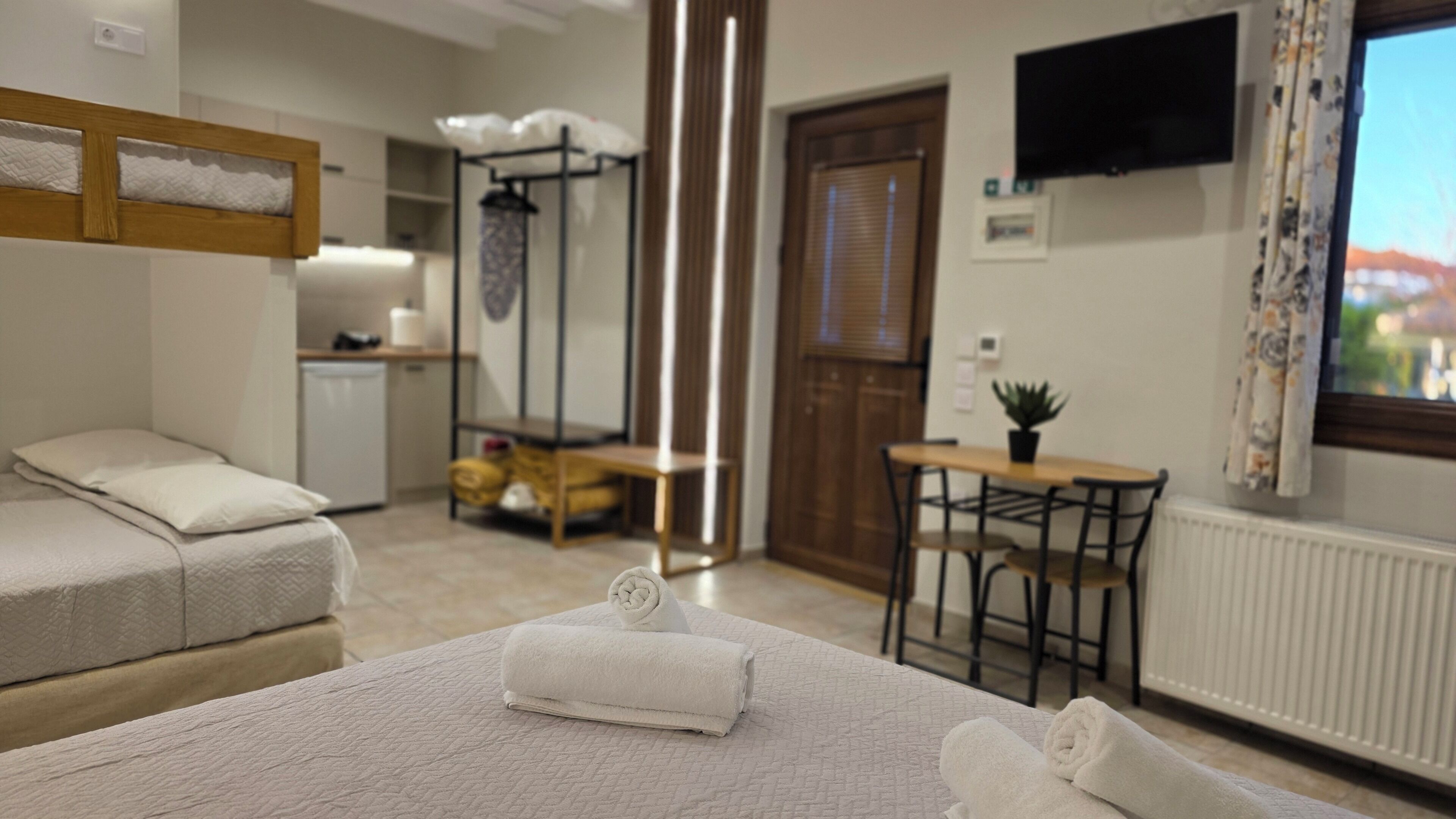 A hotel room with a bed, a small table with a plant, a TV mounted on the wall, and a kitchenette in the background.