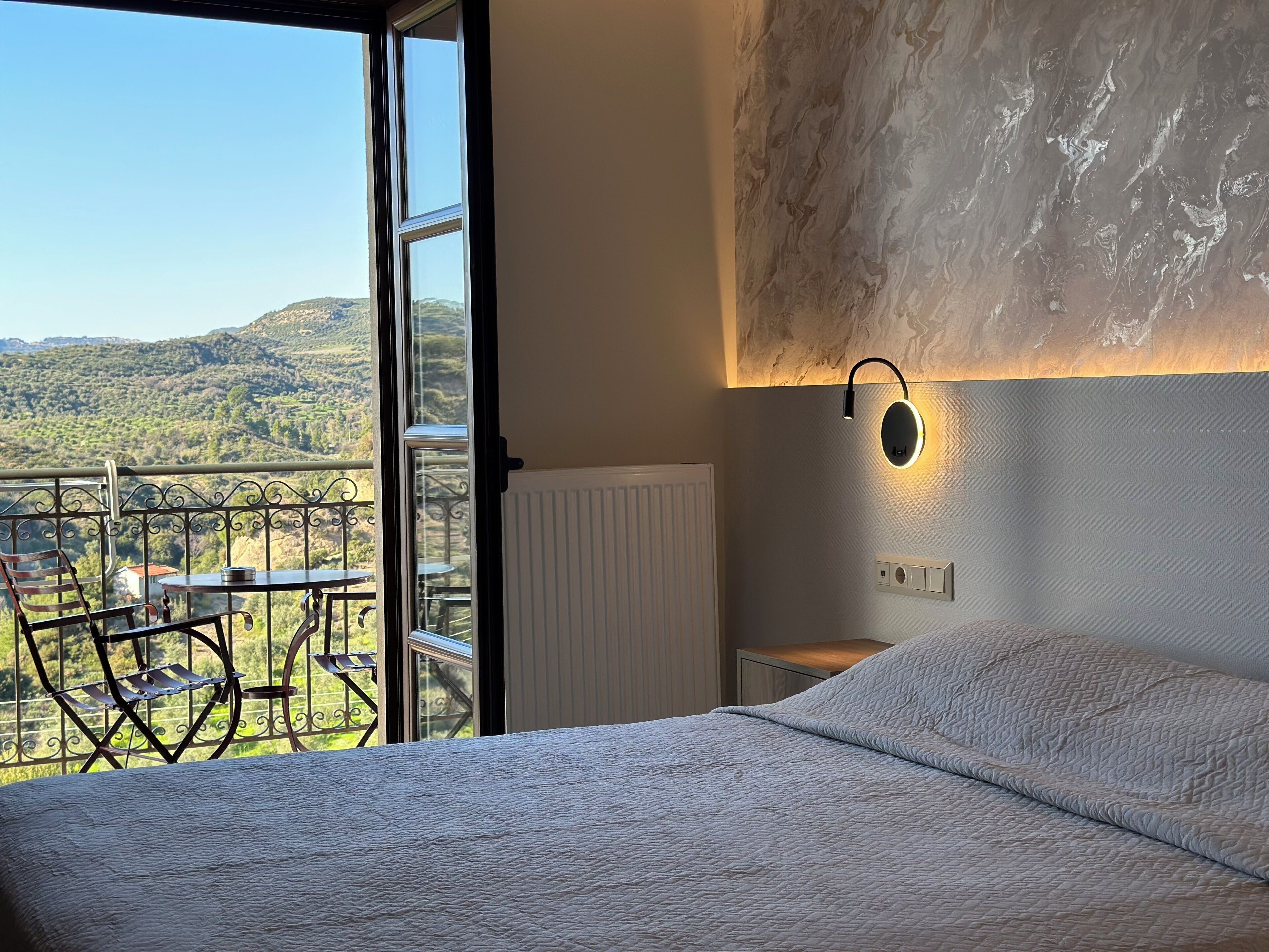 Een slaapkamer met een groot bed, een nachtkastje, een wandlamp en uitzicht op een balkon met een tafel en stoelen.