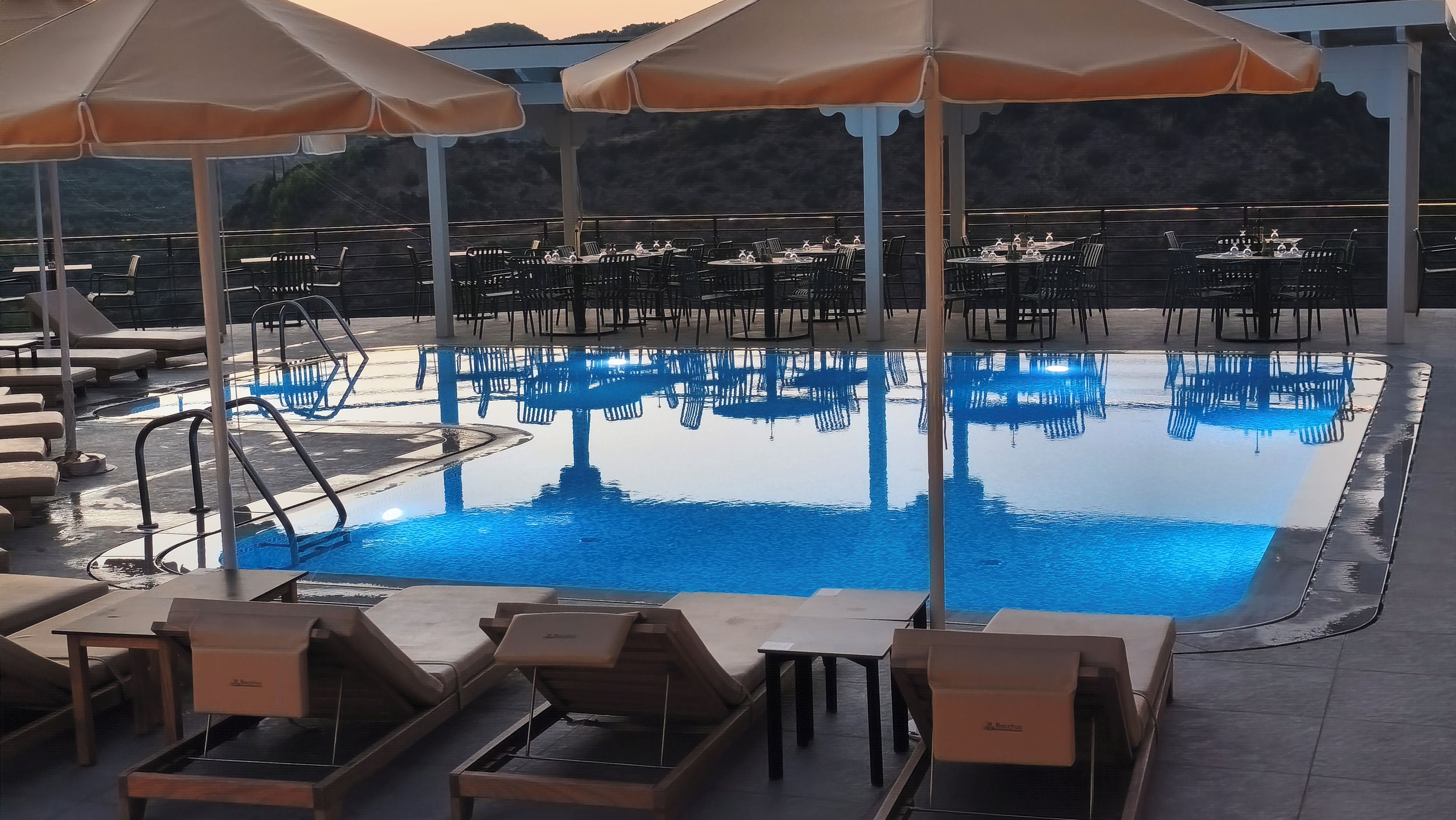 Área da piscina com espreguiçadeiras, guarda-sóis e mesas, com a piscina refletindo o céu.