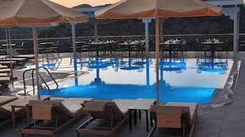 Área da piscina com espreguiçadeiras, guarda-sóis e mesas, com a piscina refletindo o céu.