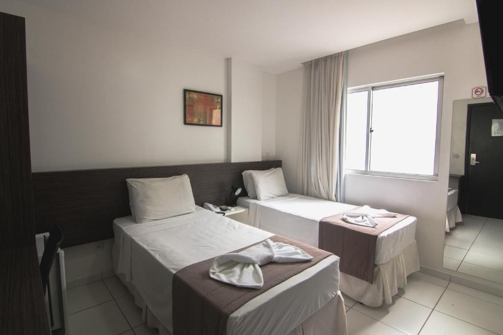 Habitación de hotel con dos camas individuales, una mesita de noche con teléfono, una ventana con cortinas y un cuadro en la pared.