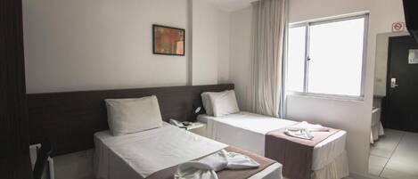Habitación de hotel con dos camas individuales, una mesita de noche con teléfono, una ventana con cortinas y un cuadro en la pared.
