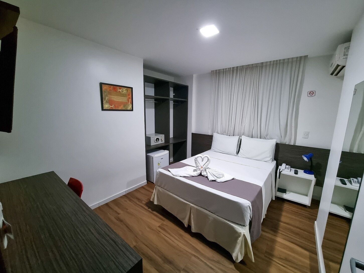 Quarto de hotel com cama, mesa de cabeceira, escrivaninha, guarda-roupa e ar condicionado.