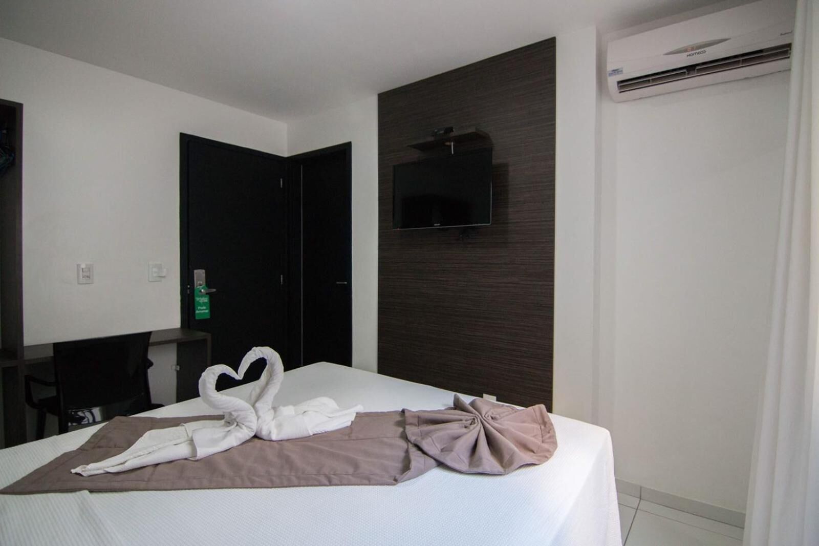 A hotel room with a bed, a flat-screen TV mounted on the wall, an air conditioning unit, and a desk with a chair.