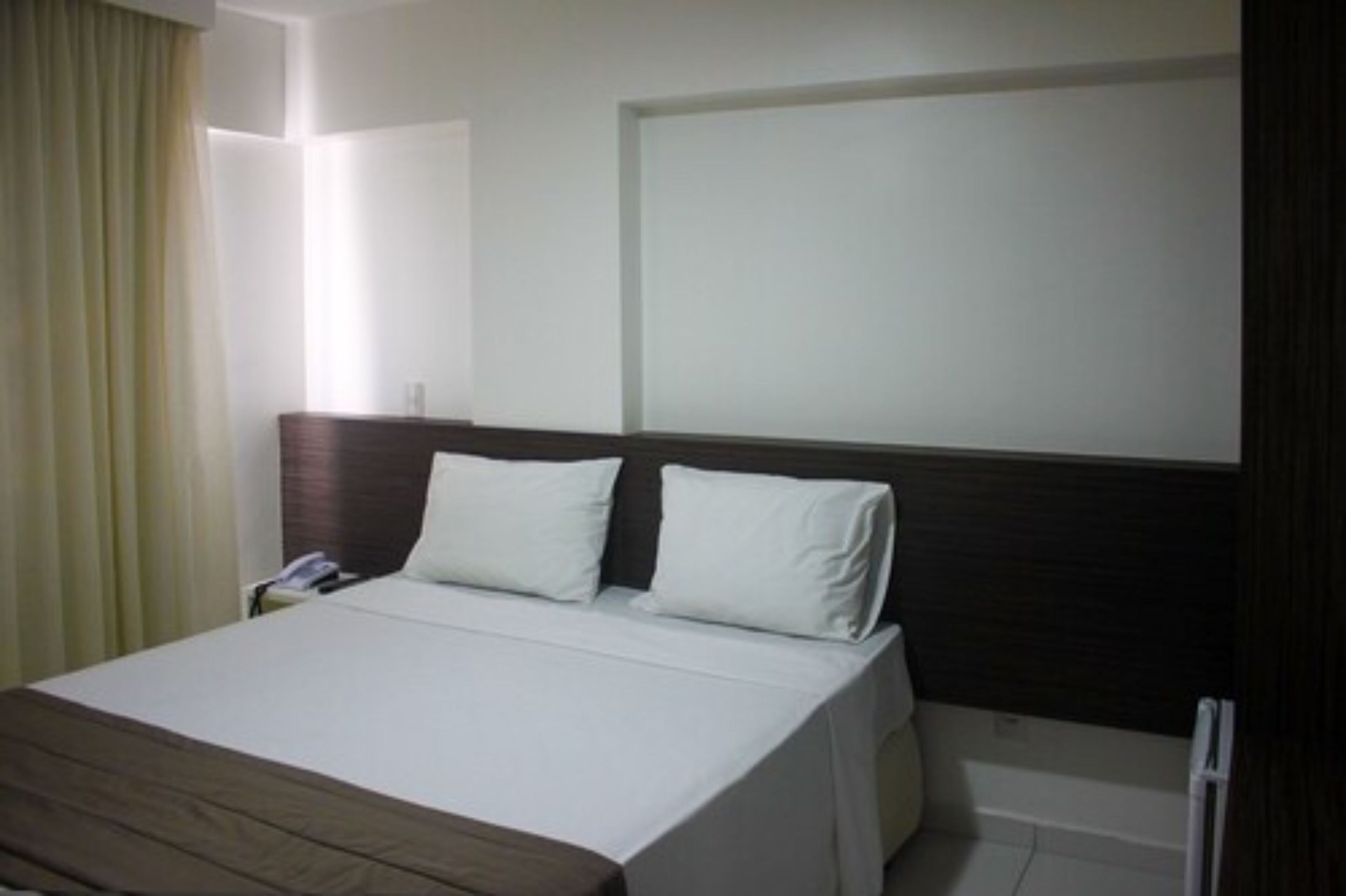 Quarto de hotel com cama, mesa de cabeceira e janela com cortinas.