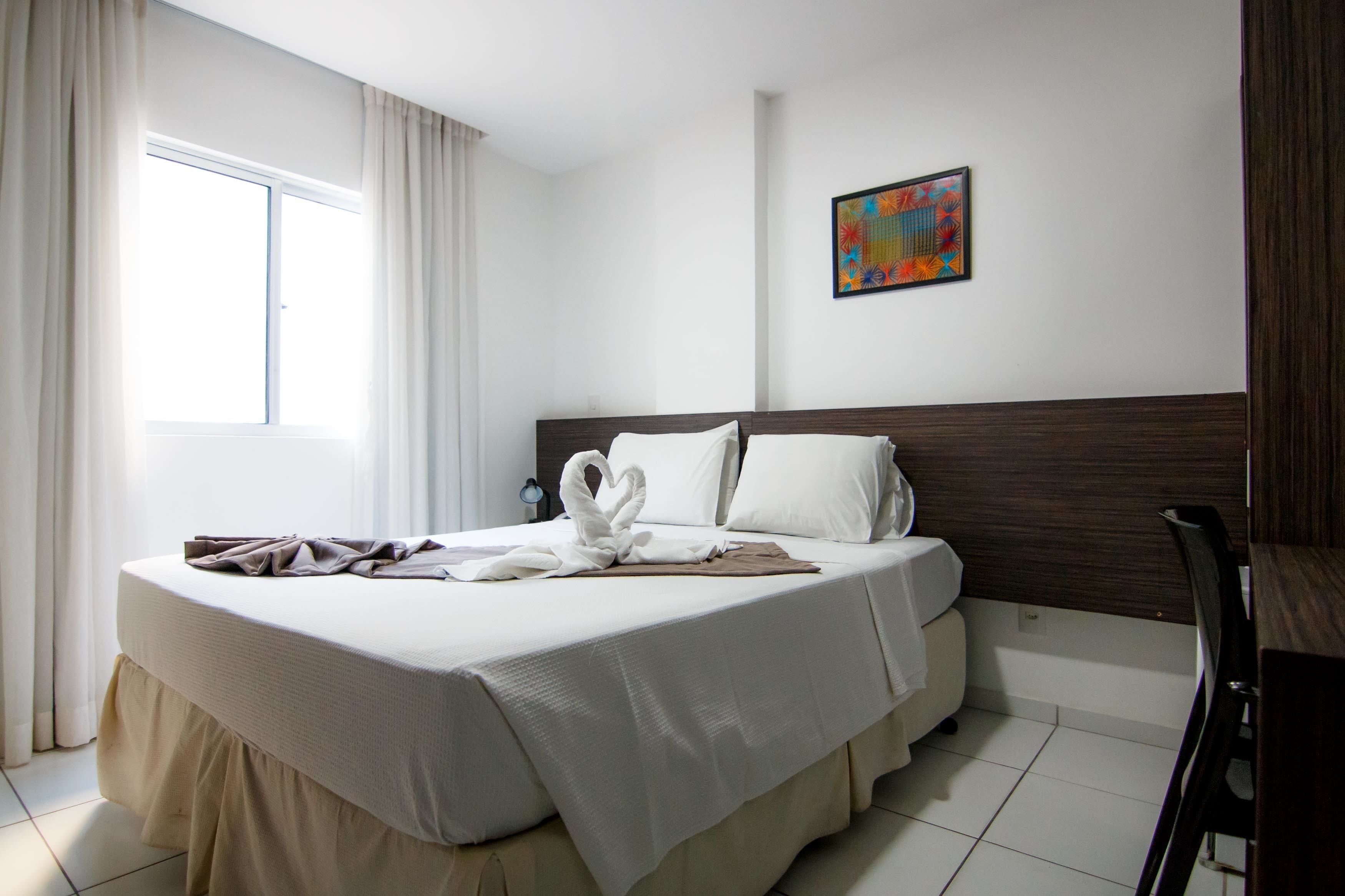 A hotel room with a large bed, a headboard, a bedside table, a chair, a window with curtains, and a colorful wall decoration.