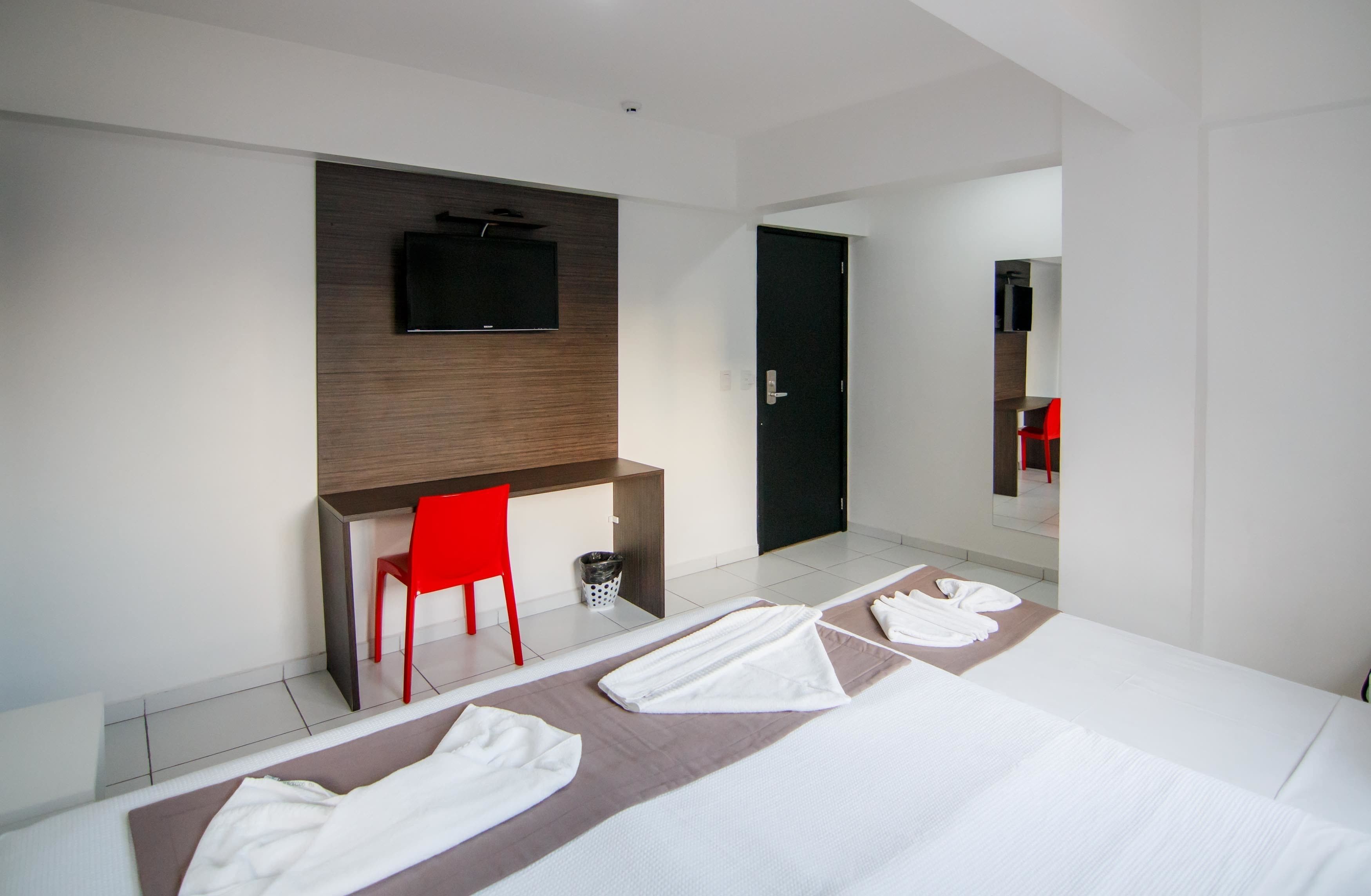 A modern hotel room with a bed, a desk, a red chair, and a TV mounted on the wall.