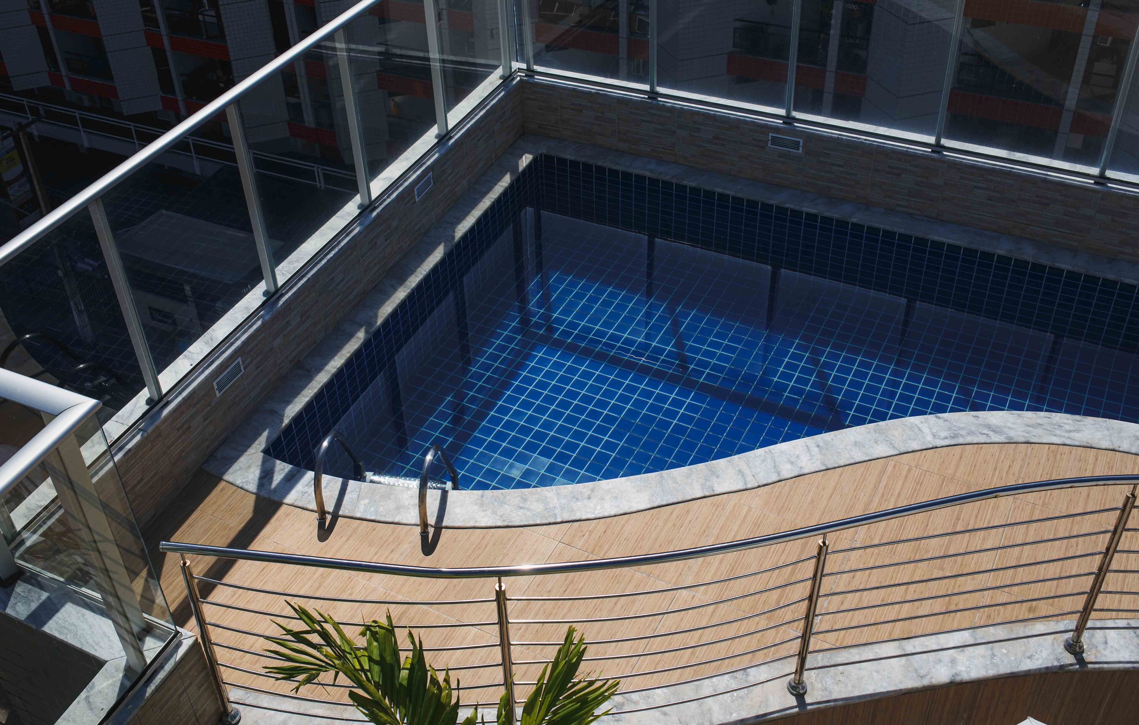 Una piscina en la azotea con baldosas azules cristalinas, rodeada de barandillas de vidrio y una terraza de madera.