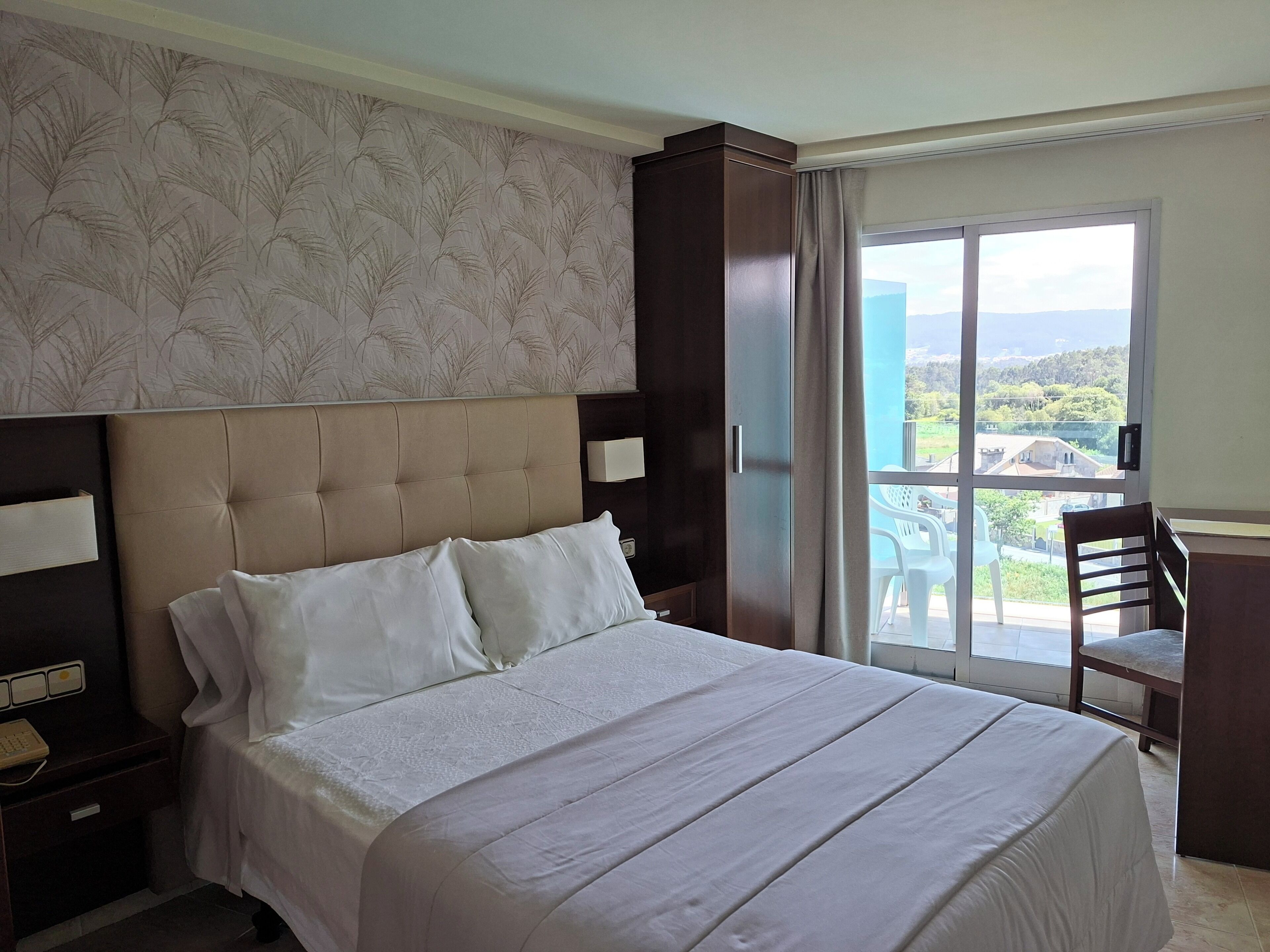 A hotel room with a large bed, a nightstand, a desk, and a view of the outdoors.