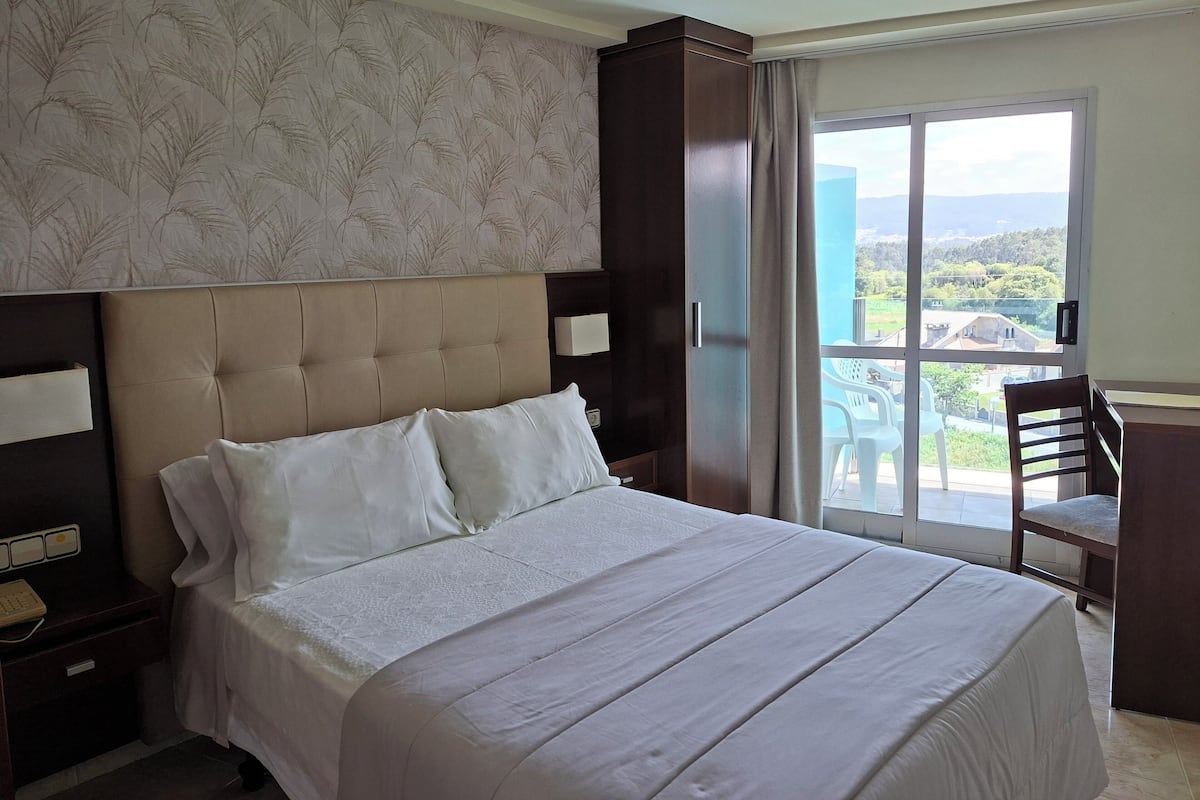 A hotel room with a large bed, a nightstand, a desk, and a view of the outdoors.