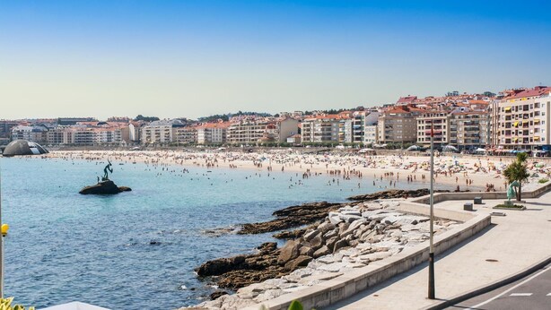 Paisagem costeira com praia, linha de costa rochosa e edifícios ao fundo.