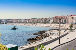 A coastal cityscape with a beach, rocky shoreline, and buildings in the background. - Troncoso (Sanxenxo)