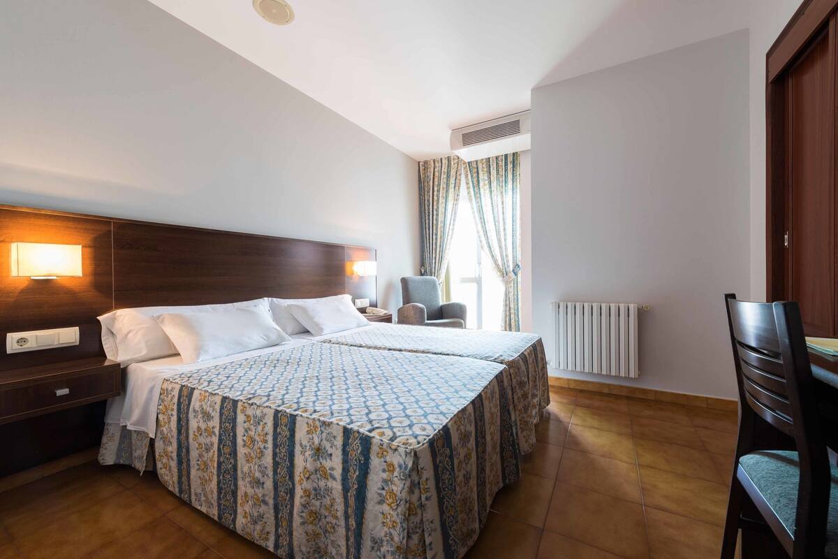 A hotel room with a large bed, a desk, a chair, and a window with curtains.
