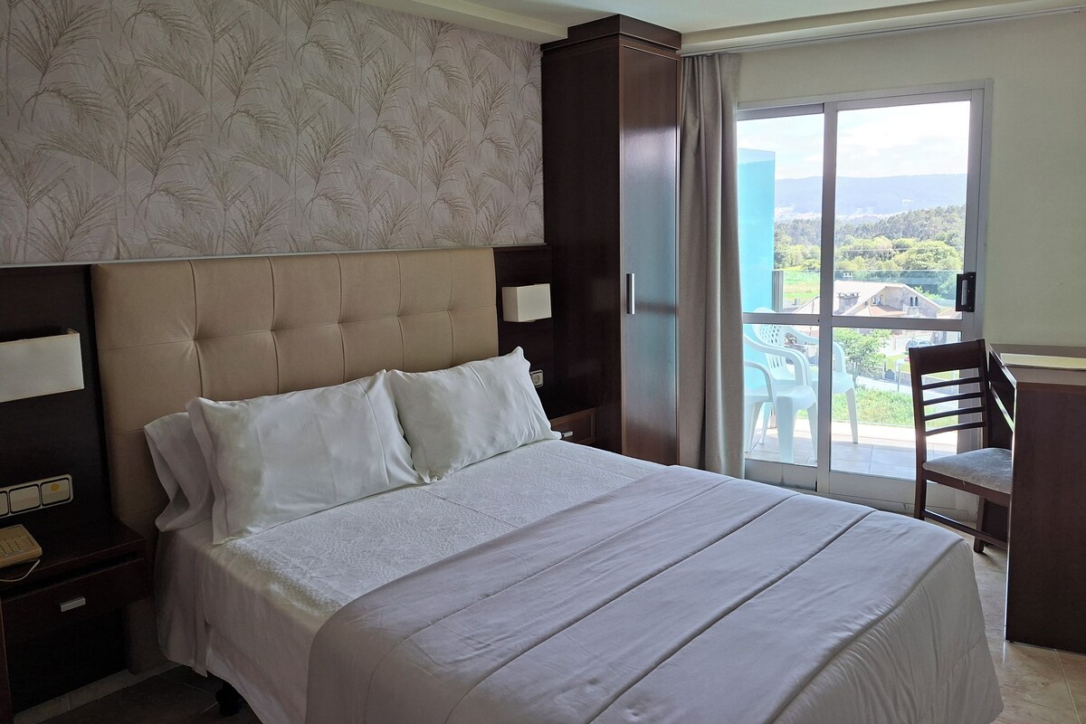 A hotel room with a large bed, a nightstand, a desk, and a view of the outdoors.
