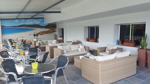 Área de estar coberta ao ar livre, com mobiliário de vime e um mural retratando uma cena de praia.