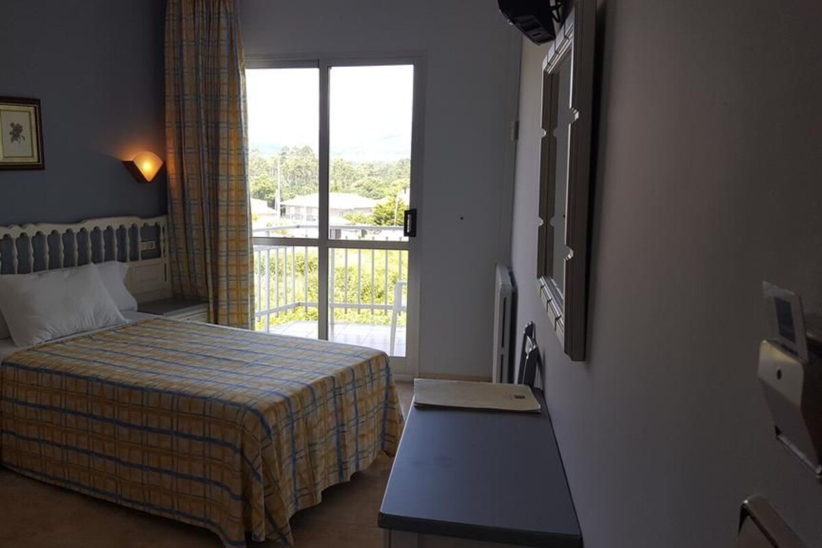 A bedroom with a bed, a nightstand, a desk, and a window with a view of greenery.
