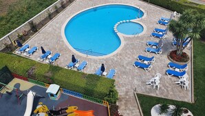 Piscina com cadeiras de descanso azuis e brancas, área de recreação infantil e um pequeno jardim com palmeiras.