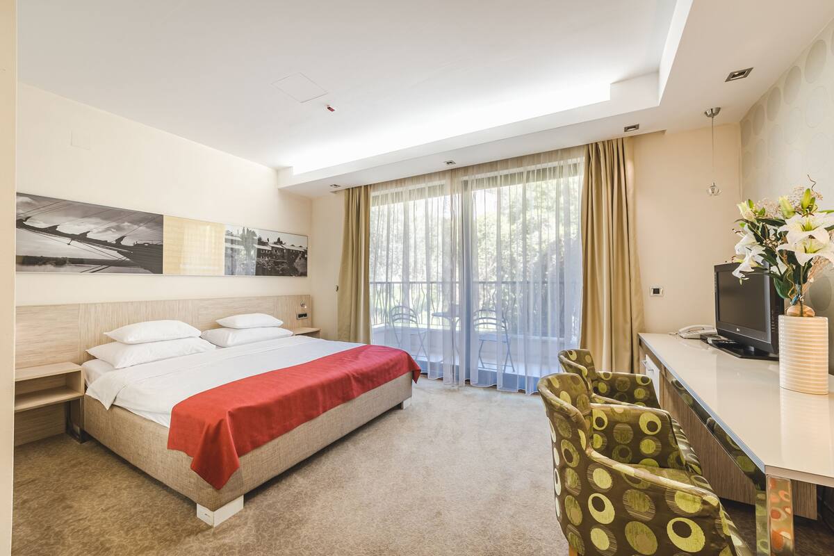Habitación de hotel con cama, escritorio, televisor, silla y ventana con cortinas.