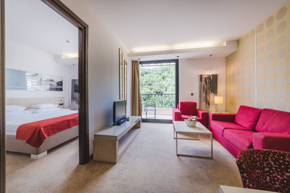 Habitación de hotel con cama, televisor, sofá rojo, mesa de centro y balcón con vistas a zonas verdes.