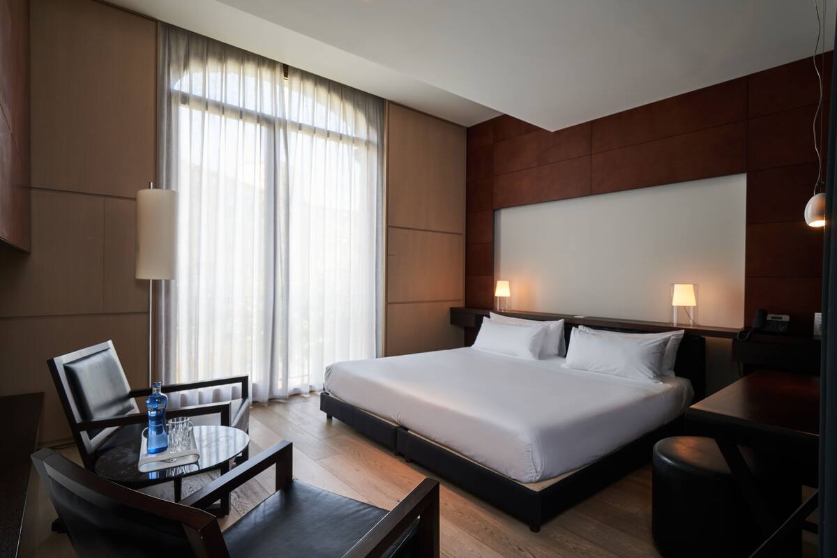 Ein Hotelzimmer mit Bett, Nachttischen, Schreibtisch, Stuhl, Lampe und einem fensterbelagten Fenster.
