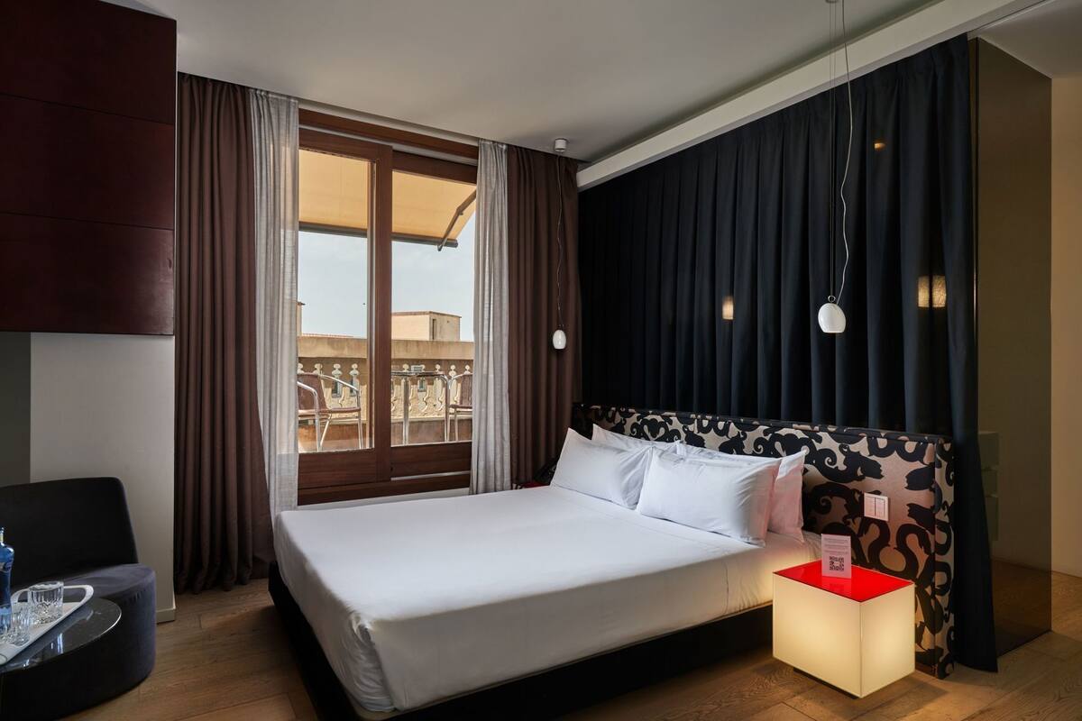 Ein Hotelzimmer mit Bett, einem Sessel, einem Nachttisch mit Lampe und einem Fenster mit Vorhängen.