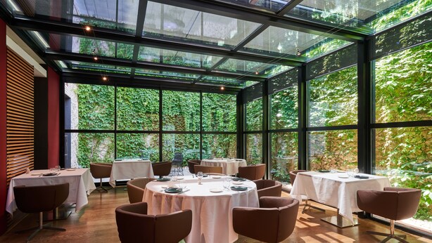 Un restaurant avec des tables rondes et des chaises marron, de grandes fenêtres et une vue sur la verdure environnante.