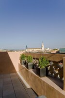 Una terraza en la azotea con plantas en macetas, cielo despejado y edificios a lo lejos, incluyendo una torre.