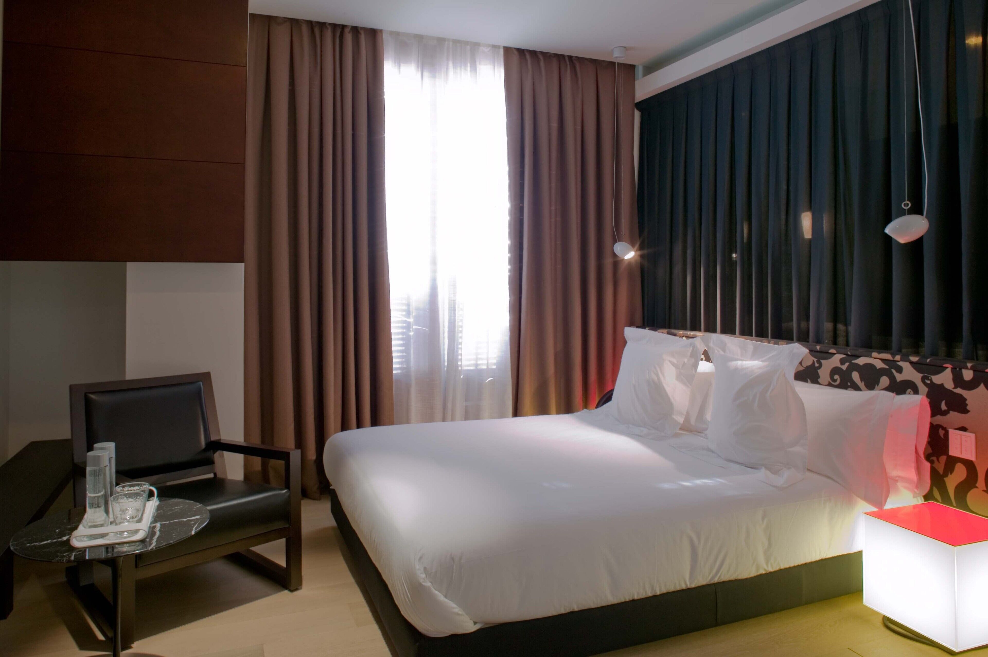 A hotel room with a bed, a chair, a table with glasses and a bottle, and a nightstand with a lamp.